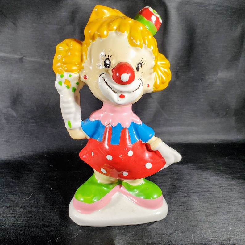 Pu&ograve; includere: Statuetta di clown in ceramica con capelli gialli, naso rosso e abito colorato. Il clown indossa un vestito rosso a pois bianchi, un top blu e scarpe verdi. Tiene un guanto bianco con un oggetto verde e giallo.