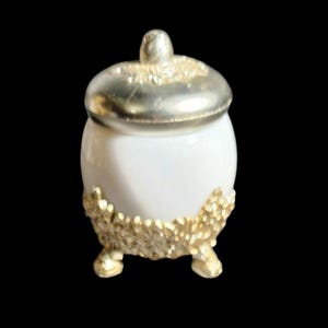 May include: A small, white ceramic jar with a silver lid and gold-colored accents. The jar has a rounded shape and sits on three small feet. The lid has a small knob on top.