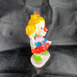 Pu&ograve; includere: Statuetta di clown in ceramica con capelli gialli, naso rosso e vestito rosso a pois bianchi. Il clown tiene in mano un fiore giallo e si trova su una base bianca. La statuetta ha un viso dipinto con un'espressione sorridente.