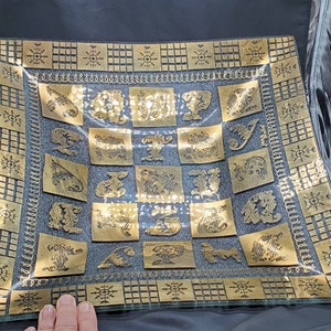 May include: A square glass serving tray with a gold-colored design. The tray has a geometric pattern around the edge and a repeating pattern of stylized floral designs in the center.
