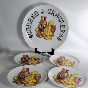 May include: A set of five white ceramic plates with a cheese and cracker theme. The large plate has the words "Cheese & Crackers" printed on it. The smaller plates feature a depiction of cheese, wine, grapes, and crackers.