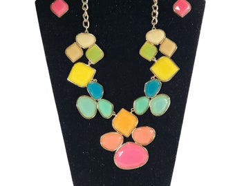 May include: A colorful multi-gemstone necklace and matching earrings set. The necklace features a gold chain with a variety of pastel-colored stones in different shapes and sizes. The earrings are small and feature a single pink stone.