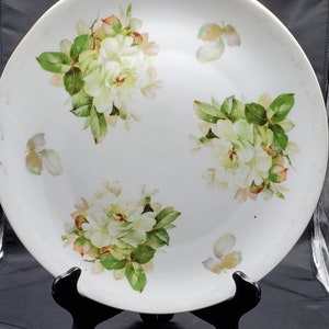 May include: A white porcelain plate with a floral design. The plate features three clusters of white roses with green leaves and pink accents. The plate is on a black stand.