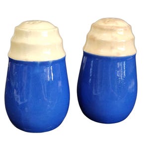 Op de afbeelding: Een paar keramische zout- en peperstrooiers. De strooiers zijn blauw met een crèmekleurige bovenkant. De bovenkanten hebben een geribbeld ontwerp en kleine gaatjes om de inhoud te verdelen. De strooiers hebben een glanzende afwerking en een klassieke, vintage uitstraling.
