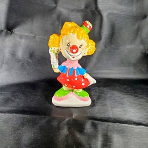 Pu&ograve; includere: Una statuetta di clown in ceramica con capelli gialli, naso rosso e cappello colorato. Il clown indossa un colletto rosa, un top blu e un vestito rosso a pois bianchi. Il clown tiene in mano un cono gelato colorato.
