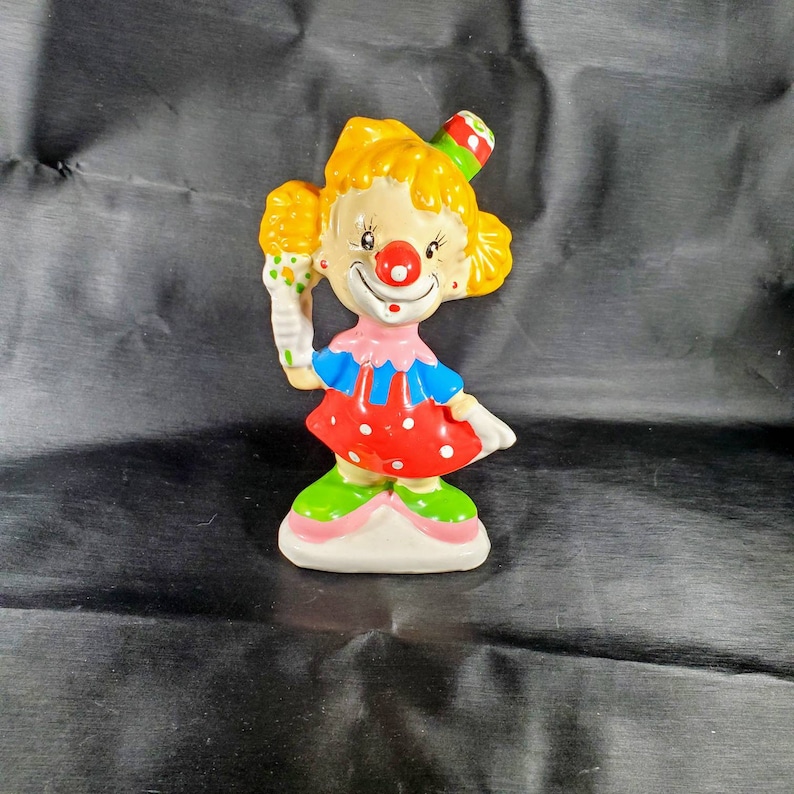 Pu&ograve; includere: Una statuetta di clown in ceramica con capelli gialli, naso rosso e cappello colorato. Il clown indossa un colletto rosa, un top blu e un vestito rosso a pois bianchi. Il clown tiene in mano un cono gelato colorato.