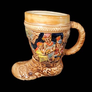 May include: A brown ceramic boot-shaped beer stein with a handle. The stein is decorated with a scene of three people in traditional German clothing playing music.