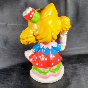 Pu&ograve; includere: Statuina vintage di una ragazza con i capelli gialli, che indossa un top blu, un vestito rosso a pois e un cappello da festa. La statuina ha una base bianca con dettagli rosa e verdi. La figura tiene in mano un oggetto bianco.