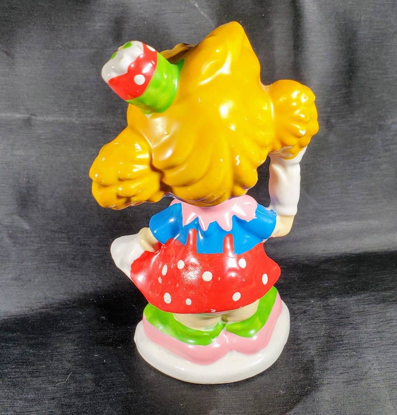 Pu&ograve; includere: Statuina vintage di una ragazza con i capelli gialli, che indossa un top blu, un vestito rosso a pois e un cappello da festa. La statuina ha una base bianca con dettagli rosa e verdi. La figura tiene in mano un oggetto bianco.