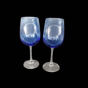 Puede incluir: Dos copas de vino de vidrio transparente con bases azules. Cada copa tiene un diseño de concha marina grabado en blanco.