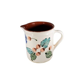 May include: A white ceramic pitcher with a brown rim and a floral design featuring blue, green, and orange colors. The pitcher has a handle and is perfect for serving beverages.
