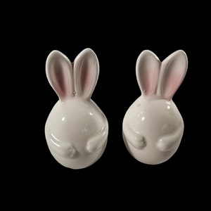 May include: A pair of white ceramic bunny salt and pepper shakers with pink ears. The bunnies are sitting upright with their paws crossed.