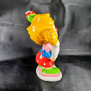 Pu&ograve; includere: Una statuetta di clown in ceramica con capelli gialli, naso rosso e un abito colorato. Il clown tiene in mano un lecca-lecca verde e rosso. La statuetta ha una base bianca con accenti rosa e verdi. L'abito del clown comprende un top rosa, sezioni blu e rosse e scarpe verdi.