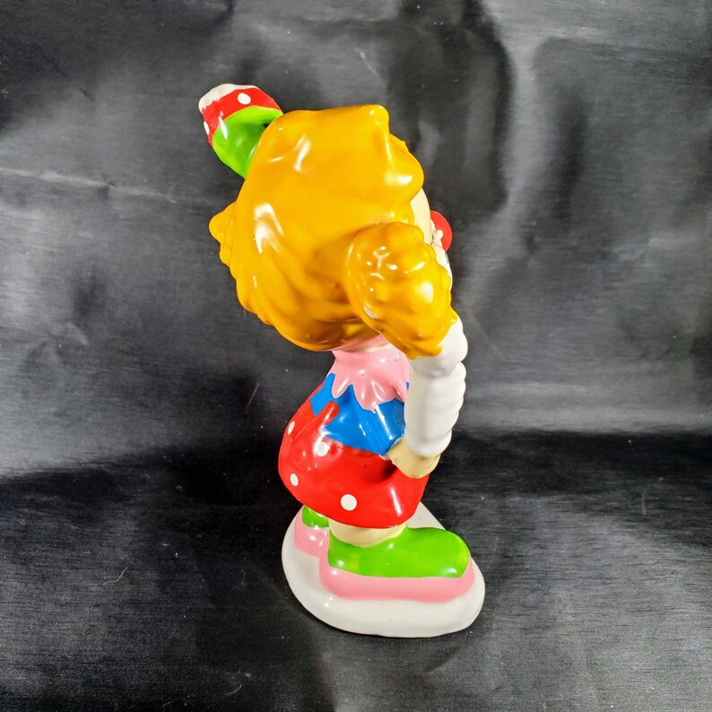 Pu&ograve; includere: Una statuetta di clown in ceramica con capelli gialli, naso rosso e un abito colorato. Il clown tiene in mano un lecca-lecca verde e rosso. La statuetta ha una base bianca con accenti rosa e verdi. L'abito del clown comprende un top rosa, sezioni blu e rosse e scarpe verdi.
