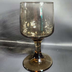 Op de afbeelding: Een helder glazen wijnglas met een bruin getinte voet en steel. Het glas heeft een ronde kom en een brede voet.