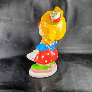 Pu&ograve; includere: Statuina vintage in ceramica di una bambina con capelli gialli, che indossa un vestito rosso a pois, un top blu e scarpe bianche. La statuina ha un piccolo cupcake sulla testa e poggia su una base bianca, rosa e verde. Oggetto da collezione.