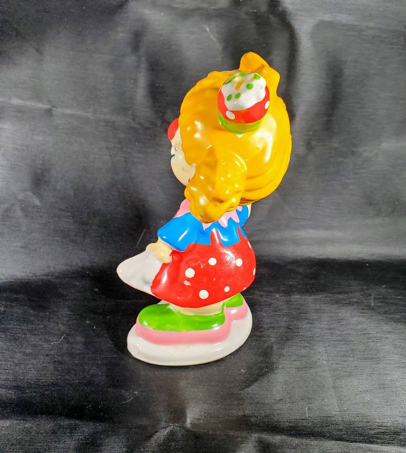 Pu&ograve; includere: Statuina vintage in ceramica di una bambina con capelli gialli, che indossa un vestito rosso a pois, un top blu e scarpe bianche. La statuina ha un piccolo cupcake sulla testa e poggia su una base bianca, rosa e verde. Oggetto da collezione.