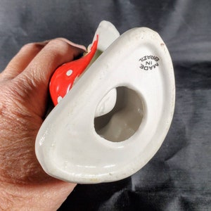 Pu&ograve; includere: Una statuetta in ceramica bianca con un elemento rosso a pois. La statuetta ha una forma curva con un foro al centro. Le parole "MADE IN BRAZIL" sono stampate sulla base.