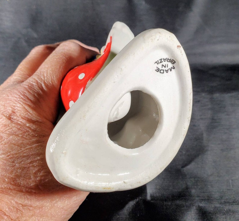 Pu&ograve; includere: Una statuetta in ceramica bianca con un elemento rosso a pois. La statuetta ha una forma curva con un foro al centro. Le parole "MADE IN BRAZIL" sono stampate sulla base.