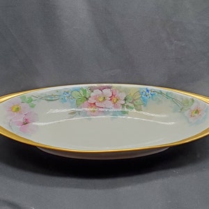 May include: A white oval serving dish with a gold rim and floral design. The dish has two handles on either side. The flowers are pink, blue, and green.