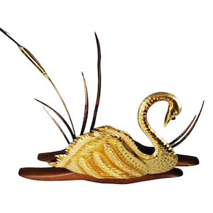 May include: A gold swan sculpture with a detailed, textured design, perched on a wooden base with a single, thin, gold stem extending upwards.