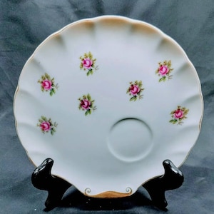 May include: A white porcelain plate with a scalloped edge and a gold rim. The plate has a small, round indentation in the centre and is decorated with pink roses.