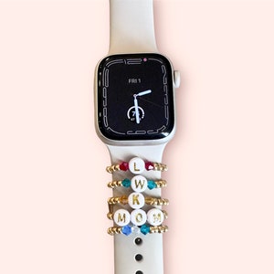 May include: A white Apple Watch with a black face displaying "FRI 1" on the screen. The watch is adorned with five gold beaded bracelets with white letter beads spelling out "LWK MOM". The beads are interspersed with colorful crystal beads.