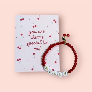 May include: A red beaded bracelet with the name "MOLLY" spelled out in white letter beads. The bracelet has a cherry charm and is next to a card that says "you are cherry special to me!" with cherry illustrations.