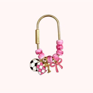 May include: Gold-toned keychain with pink beads and charms. Charms include a soccer ball, a pink and white checkered heart, the number 1, and a pink bow. The keychain is a decorative accessory.