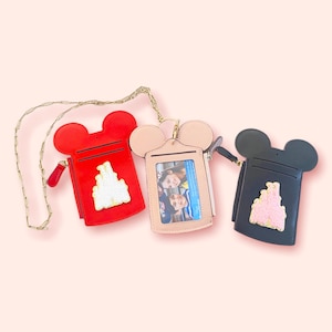 May include: Three Mickey Mouse-shaped ID card holders with gold chains. The holders are red, pink, and black. Each holder has a gold castle design and a clear plastic window for a photo.