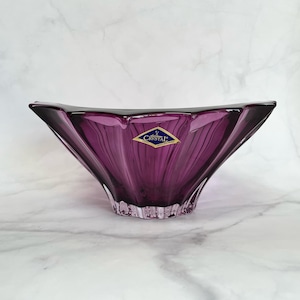 May include: A purple glass bowl with a starburst design. The bowl has a label that reads "Bohemian Crystal".
