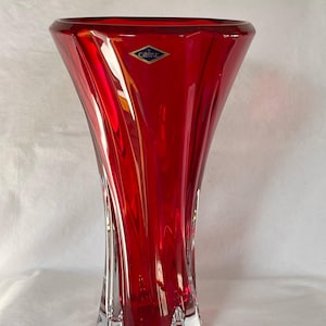 Könnte beinhalten: Eine hohe, rote Kristallvase mit ausgestelltem Rand und klarer, strukturierter Basis. Die Vase hat eine glänzende Oberfläche und ein Etikett mit der Aufschrift "Crystal". Das Design weist vertikale, geschwungene Linien auf.