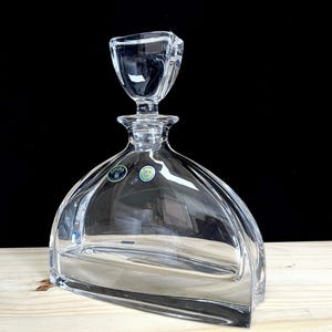 May include: A clear glass decanter with a square-shaped stopper. The decanter has a wide base and a narrow neck. The decanter is made of crystal and has a label that says "BOHEMIA 24% PbO Crystal".