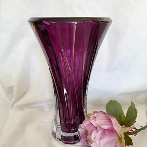 Könnte beinhalten: Eine hohe, amethystfarbene Glasvase mit ausgestelltem Rand und facettiertem Boden. Die Vase hat vertikale, wirbelnde Rillen. Eine rosa Pfingstrose mit grünen Blättern liegt neben der Vase.