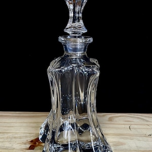 Pode incluir: Uma jarra de vidro transparente com uma rolha decorativa. A jarra tem uma base alargada e um pescoço alto e fino. A rolha é ornamentada, com um design floral. A jarra está sobre uma superfície de madeira.