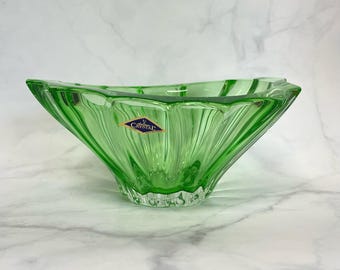 May include: A green glass bowl with a unique, angular design. The bowl has a flared shape with vertical ridges and a clear base. A blue and gold sticker with the word "Crystal" is affixed to the side.