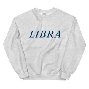 Könnte beinhalten: Ein hellgraues Sweatshirt mit Rundhalsausschnitt und dem Wort LIBRA, das in einer dunkelblauen Serifenschrift auf der Brust gedruckt ist. Das Kleidungsstück hat lange Ärmel, einen gerippten Kragen und gerippte Bündchen aus meliertem Stoff.