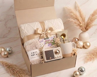 May include: A gift box containing a cream-colored cable knit blanket tied with a gold ribbon, a pair of beige fuzzy socks labeled COZY SOCKS, a small vintage-style radio, a ceramic mug with a gold spoon, and a packet labeled HOT CACAO.