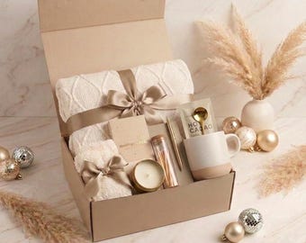 May include: A gift box containing a cream-colored cable knit blanket tied with a beige ribbon, a small fuzzy item with a bow, a candle in a gold tin, a glass vial of matches, a gold pen, a notebook, a mug, and a gold spoon with hot cacao mix.