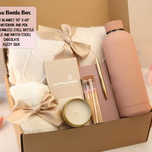 Puede incluir: Una caja de regalo con una manta acogedora, un cuaderno, un bol&iacute;grafo, una botella de acero inoxidable, una vela, cerillas y un calcet&iacute;n suave. La manta mide 127 cm x 152 cm y la botella 500 ml. La caja es de color rosa y beige.