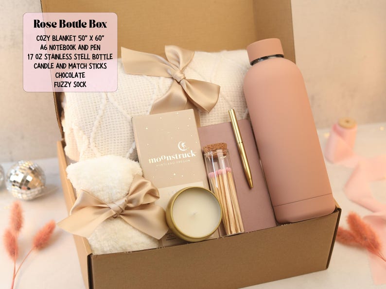 Puede incluir: Una caja de regalo con una manta acogedora, un cuaderno, un bol&iacute;grafo, una botella de acero inoxidable, una vela, cerillas y un calcet&iacute;n suave. La manta mide 127 cm x 152 cm y la botella 500 ml. La caja es de color rosa y beige.