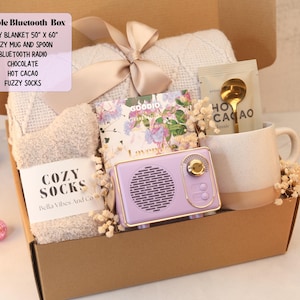 May include: A gift box containing a 50" x 60" blanket with a ribbon, fuzzy socks labeled "COZY SOCKS", a Goodio chocolate bar, a packet of hot cacao with a gold spoon, and a purple Bluetooth radio next to a speckled ceramic mug.