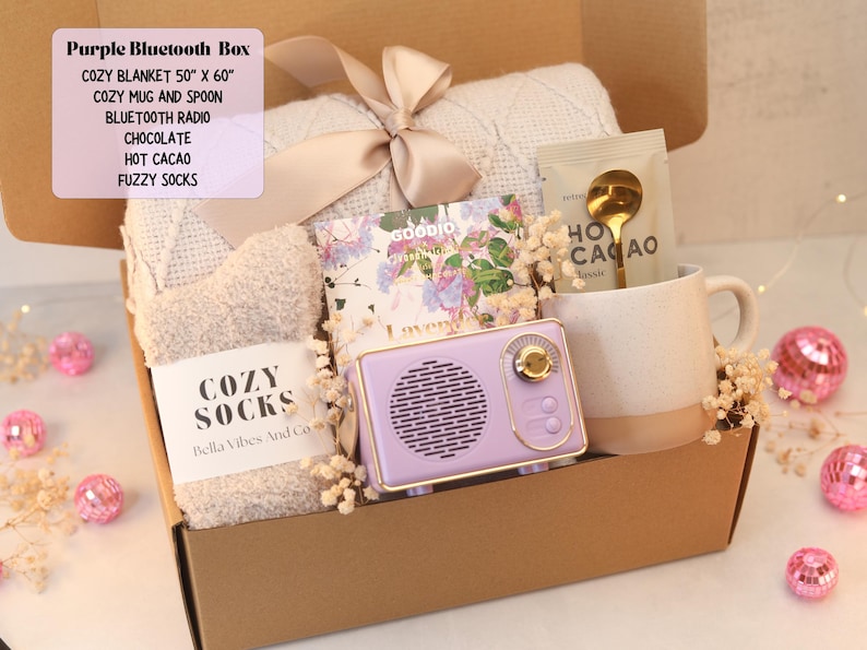 May include: A gift box containing a 50" x 60" blanket with a ribbon, fuzzy socks labeled "COZY SOCKS", a Goodio chocolate bar, a packet of hot cacao with a gold spoon, and a purple Bluetooth radio next to a speckled ceramic mug.