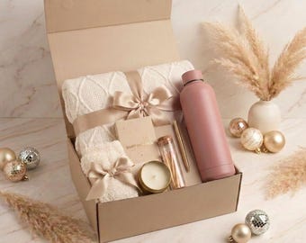Puede incluir: Una caja de regalo que contiene una manta de punto trenzado color crema atada con una cinta color champán, una botella de agua rosa polvoriento, un pequeño cuaderno con la etiqueta 'moments', un bolígrafo dorado, un vial de cristal con cerillas y una vela.