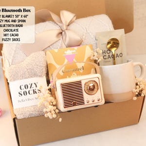 May include: A gift box containing a beige knit blanket with a ribbon, a pair of fuzzy socks, a Goodio caramel chocolate bar, a speckled ceramic mug with a gold spoon, a packet of hot cacao, and a vintage-style beige Bluetooth radio speaker.