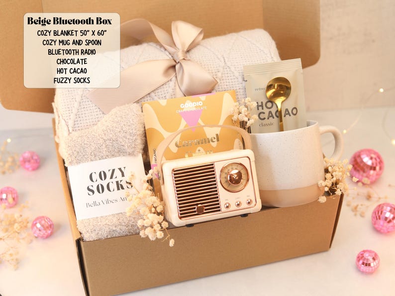 May include: A gift box containing a beige knit blanket with a ribbon, a pair of fuzzy socks, a Goodio caramel chocolate bar, a speckled ceramic mug with a gold spoon, a packet of hot cacao, and a vintage-style beige Bluetooth radio speaker.