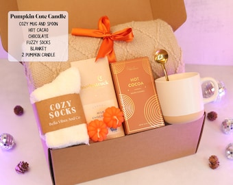 Op de afbeelding: Een geschenkdoos met een beige kabelgebreide deken vastgebonden met een oranje lint, witte pluizige sokken, een reep melkchocolade, een doos warme chocolademelk, twee oranje pompoenvormige kaarsen en een witte mok met een gouden lepel.