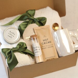 Puede incluir: Caja de regalo con artículos de cuidado personal. Incluye una toalla blanca, una vela con el texto "bella vibes only", cacao caliente, cerillas, una taza y una cuchara dorada. La caja está adornada con lazos de satén verde.