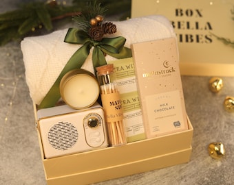 May include: A gift box containing a white cable-knit blanket tied with a green ribbon and pinecones, a gold-toned candle, a glass vial of matches, a green tea packet, and a Moonstruck milk chocolate bar next to a small cream-colored radio.