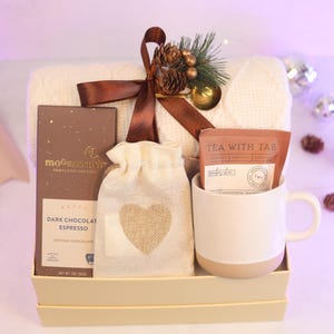 May include: A gift set in a box containing a cream-colored cable knit blanket tied with a brown ribbon, a bar of Moonstruck dark chocolate espresso, a small beige drawstring pouch with a burlap heart, and a white ceramic mug with a tea packet.
